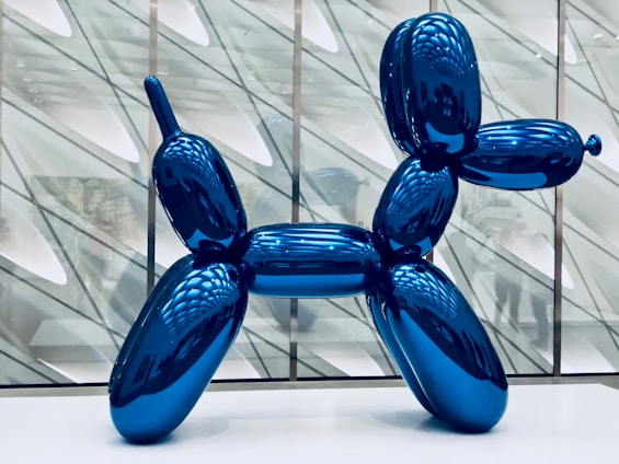 A large, shiny blue sculpture resembling a balloon animal in the shape of a dog stands prominently in a modern gallery space. The background consists of geometric patterns and reflected light, creating a sense of depth.