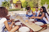 A group of friends are seated around a low outdoor table decorated with plates, glassware, and snacks. The setting appears casual and summery, with lush greenery and large yellow balloons in the background. The friends are engaged in conversation and smiling, giving off a relaxed and joyful atmosphere.