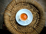 A cup of tea with a rich brown color sits in a white saucer on top of a woven circular mat made from natural fibers.