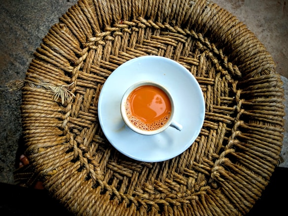 A cup of tea with a rich brown color sits in a white saucer on top of a woven circular mat made from natural fibers.