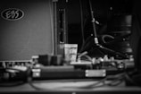 A black and white photograph featuring an amplifier with the brand 'EBS' visible, along with a small plastic cup in the center. Cables and audio equipment are scattered around, with a microphone stand and other musical gear in the background.