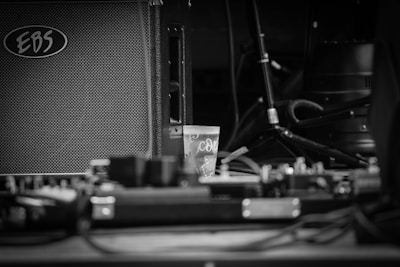 A black and white photograph featuring an amplifier with the brand 'EBS' visible, along with a small plastic cup in the center. Cables and audio equipment are scattered around, with a microphone stand and other musical gear in the background.