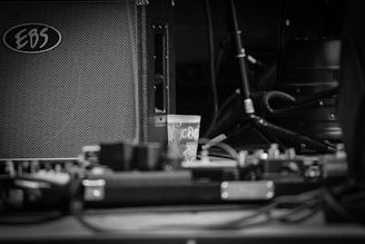A black and white photograph featuring an amplifier with the brand 'EBS' visible, along with a small plastic cup in the center. Cables and audio equipment are scattered around, with a microphone stand and other musical gear in the background.