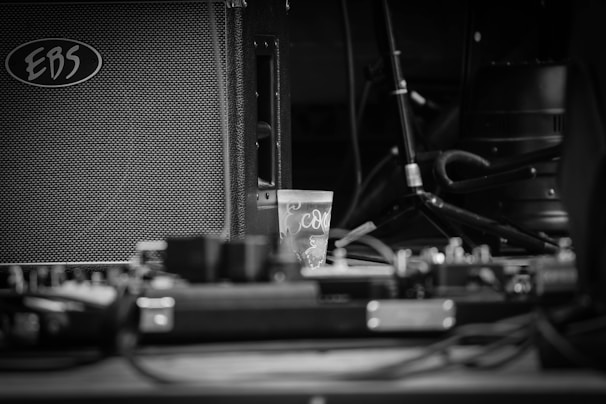 A black and white photograph featuring an amplifier with the brand 'EBS' visible, along with a small plastic cup in the center. Cables and audio equipment are scattered around, with a microphone stand and other musical gear in the background.