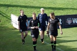 Four women in sports attire are walking across a grass field. They are wearing black uniforms with numbers and team logos. The field is green and sunny, and there is a banner in the background.