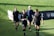 Four women in sports attire are walking across a grass field. They are wearing black uniforms with numbers and team logos. The field is green and sunny, and there is a banner in the background.