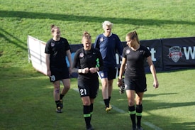 Four women in sports attire are walking across a grass field. They are wearing black uniforms with numbers and team logos. The field is green and sunny, and there is a banner in the background.