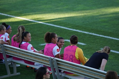 A group of female soccer players sits on a bench at the sidelines of a sports field. They are wearing sports gear, with pink bibs over their jerseys. The players appear to be enjoying themselves, with one prominently laughing. The field is well-maintained and marked with white lines.