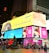 A vibrant cityscape featuring large digital billboards with advertisements. The prominent signage includes an ad for the Michelin tire on a yellow background, an AT&T ad on a blue background, and a family greeting on a pink background. There are people walking and standing around a statue in the urban environment, with bright lights illuminating the scene.