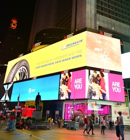 A vibrant cityscape featuring large digital billboards with advertisements. The prominent signage includes an ad for the Michelin tire on a yellow background, an AT&T ad on a blue background, and a family greeting on a pink background. There are people walking and standing around a statue in the urban environment, with bright lights illuminating the scene.