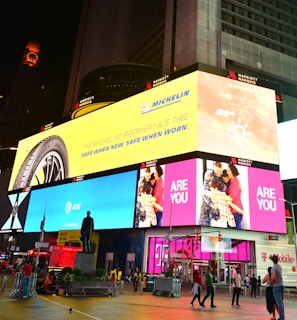 A vibrant cityscape featuring large digital billboards with advertisements. The prominent signage includes an ad for the Michelin tire on a yellow background, an AT&T ad on a blue background, and a family greeting on a pink background. There are people walking and standing around a statue in the urban environment, with bright lights illuminating the scene.