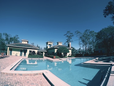 A serene outdoor swimming pool surrounded by brick pavers, adjacent to a cream-colored building featuring multiple chimneys and a gable roof. Tall trees are visible in the background under a clear blue sky, contributing to a tranquil atmosphere.