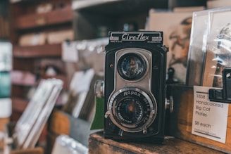 A vintage twin-lens reflex camera is prominently displayed, surrounded by various antique items. The camera has a classic design with two lenses and a distinct brand logo on top. Nearby, there are art prints and a price tag listing collectible plates for sale. The background features blurred objects, enhancing the focus on the camera.