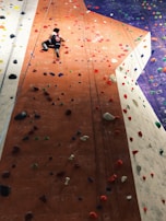 A person is climbing an indoor rock wall equipped with various colorful handholds and footholds. The wall has distinct sections in orange, white, and purple, providing a variety of paths and challenges. The climber is wearing a black outfit with a red and white design.