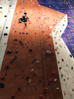 A person is climbing an indoor rock wall equipped with various colorful handholds and footholds. The wall has distinct sections in orange, white, and purple, providing a variety of paths and challenges. The climber is wearing a black outfit with a red and white design.