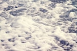 Foamy waves with a mix of white bubbles and darker water below create a textured surface, appearing as if captured at a shoreline.