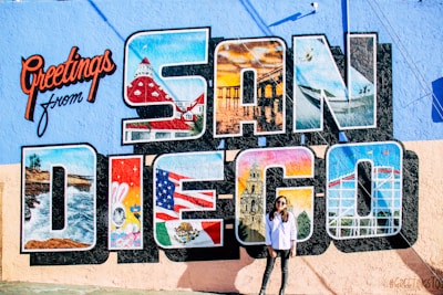 A mural features the large text 'San Diego' with various iconic images within each letter, such as a coastline, a famous building, and cultural symbols. The vibrant background also displays the words 'Greetings from' in red cursive. In front of the mural stands a person wearing sunglasses and casual clothing.