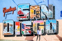 A mural features the large text 'San Diego' with various iconic images within each letter, such as a coastline, a famous building, and cultural symbols. The vibrant background also displays the words 'Greetings from' in red cursive. In front of the mural stands a person wearing sunglasses and casual clothing.