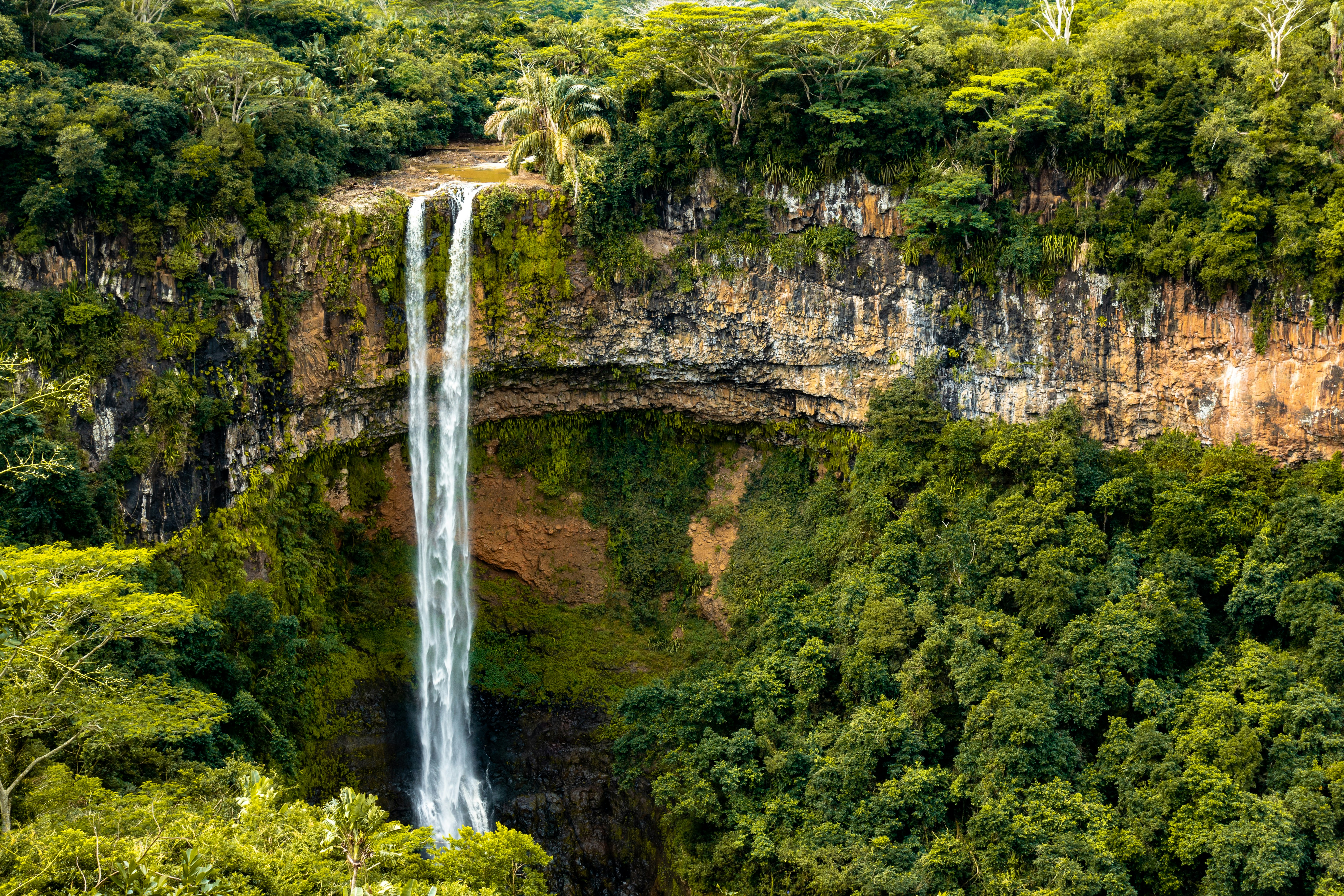 Dramatic highland waterfall cascading down mossy green cliffs surrounded by lush tropical vegetation in bright daylight