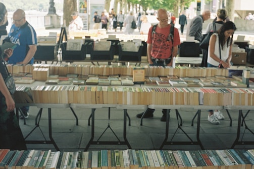 A market stall displaying a variety of books laid out on tables. Several people are browsing through the books, with a tree-lined path and other stalls visible in the background. The setting appears to be outdoors, giving a casual and relaxed atmosphere.