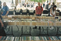 A market stall displaying a variety of books laid out on tables. Several people are browsing through the books, with a tree-lined path and other stalls visible in the background. The setting appears to be outdoors, giving a casual and relaxed atmosphere.