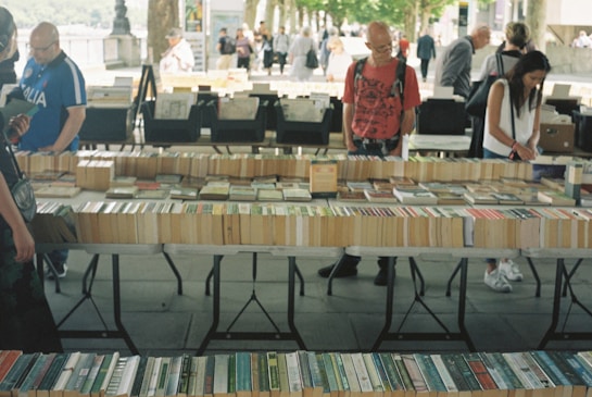 A market stall displaying a variety of books laid out on tables. Several people are browsing through the books, with a tree-lined path and other stalls visible in the background. The setting appears to be outdoors, giving a casual and relaxed atmosphere.