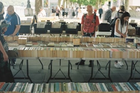 A market stall displaying a variety of books laid out on tables. Several people are browsing through the books, with a tree-lined path and other stalls visible in the background. The setting appears to be outdoors, giving a casual and relaxed atmosphere.