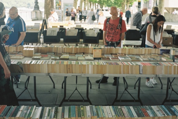 A market stall displaying a variety of books laid out on tables. Several people are browsing through the books, with a tree-lined path and other stalls visible in the background. The setting appears to be outdoors, giving a casual and relaxed atmosphere.