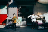 Two children stand in front of a market stall, one holding a bundle of green vegetables and the other embracing a large white stuffed bird. The scene is brightly lit by sunlight, casting strong shadows on the gray brick wall behind them. Various crates and baskets filled with produce are visible, and a red door frames the left side of the image.