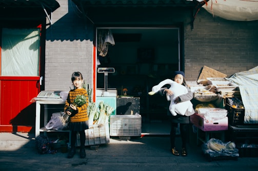 Two children stand in front of a market stall, one holding a bundle of green vegetables and the other embracing a large white stuffed bird. The scene is brightly lit by sunlight, casting strong shadows on the gray brick wall behind them. Various crates and baskets filled with produce are visible, and a red door frames the left side of the image.
