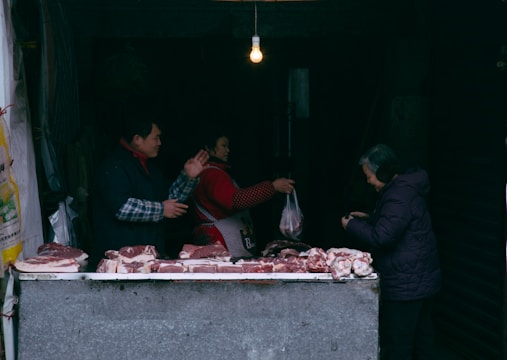 A market stall displays various cuts of raw meat on a counter. Three people are present: a vendor interacting with a customer and another person handling a plastic bag. An exposed light bulb hangs above, providing dim lighting.