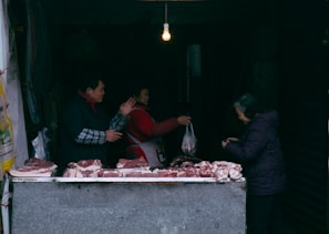 A market stall displays various cuts of raw meat on a counter. Three people are present: a vendor interacting with a customer and another person handling a plastic bag. An exposed light bulb hangs above, providing dim lighting.