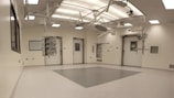 An empty, well-lit, modern operating room with white walls and ceiling. Various surgical lights and equipment are suspended from the ceiling. The room has a clean and sterile appearance with closed doors and built-in shelves.