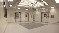 An empty, well-lit, modern operating room with white walls and ceiling. Various surgical lights and equipment are suspended from the ceiling. The room has a clean and sterile appearance with closed doors and built-in shelves.