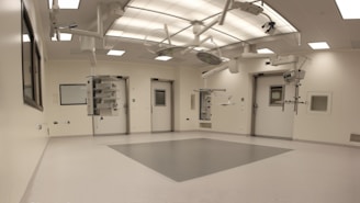 An empty, well-lit, modern operating room with white walls and ceiling. Various surgical lights and equipment are suspended from the ceiling. The room has a clean and sterile appearance with closed doors and built-in shelves.