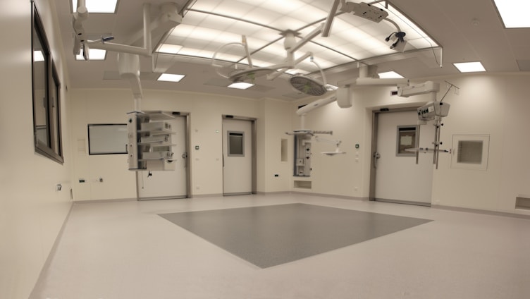 An empty, well-lit, modern operating room with white walls and ceiling. Various surgical lights and equipment are suspended from the ceiling. The room has a clean and sterile appearance with closed doors and built-in shelves.