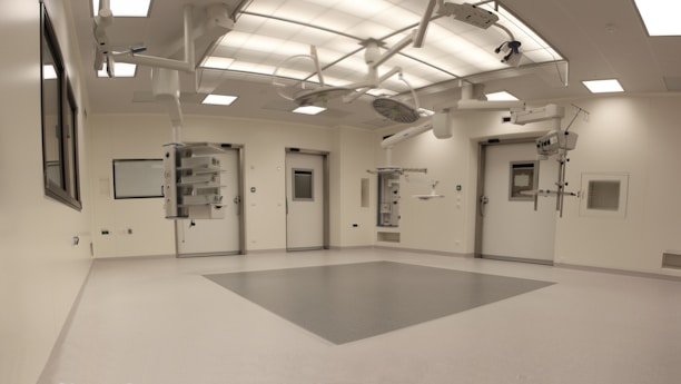 An empty, well-lit, modern operating room with white walls and ceiling. Various surgical lights and equipment are suspended from the ceiling. The room has a clean and sterile appearance with closed doors and built-in shelves.