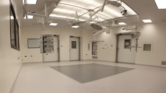 An empty, well-lit, modern operating room with white walls and ceiling. Various surgical lights and equipment are suspended from the ceiling. The room has a clean and sterile appearance with closed doors and built-in shelves.