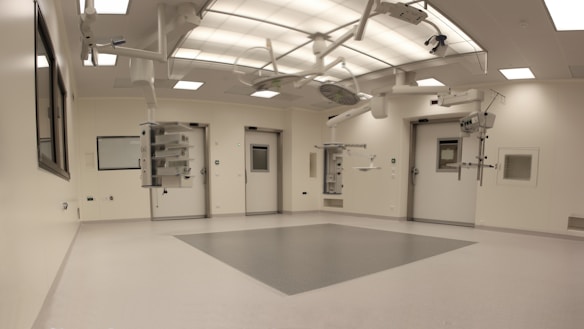 An empty, well-lit, modern operating room with white walls and ceiling. Various surgical lights and equipment are suspended from the ceiling. The room has a clean and sterile appearance with closed doors and built-in shelves.
