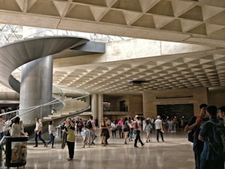 A large, modern indoor space features a prominent spiral staircase made of metal, with people walking and gathering around. The ceiling has a geometric, grid-like pattern, and the lighting is natural, filtering through large openings. Signage indicates the location is a museum or cultural institution.