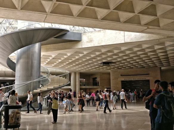 A large, modern indoor space features a prominent spiral staircase made of metal, with people walking and gathering around. The ceiling has a geometric, grid-like pattern, and the lighting is natural, filtering through large openings. Signage indicates the location is a museum or cultural institution.