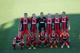 A group of ten female soccer players is posing on a green field. They are wearing red and blue uniforms with a logo on their chest. The players are arranged in two rows, with five standing and five kneeling in front. The lighting creates a distinct shadow across part of the field.