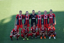 A group of ten female soccer players is posing on a green field. They are wearing red and blue uniforms with a logo on their chest. The players are arranged in two rows, with five standing and five kneeling in front. The lighting creates a distinct shadow across part of the field.