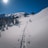 A lone skier is making their way up a snowy mountain trail under a clear blue sky. The surrounding landscape features tall, snow-covered trees and mountain peaks. The sun is shining brightly, creating long shadows and reflecting light off the pristine snow.