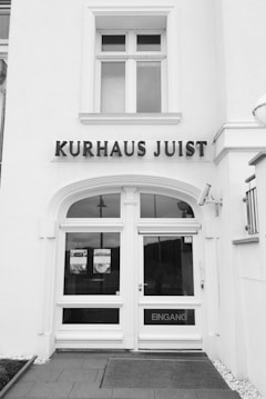 A black and white image of a building entrance with the words 'KURHAUS JUIST' prominently displayed above the door. The door is framed with large windows, sporting a sign with the word 'EINGANG', which means entrance. The facade is clean and exhibits classic architectural elements with a symmetrical design.