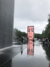 A large video screen displays a close-up of a face with closed eyes, appearing to be spraying water from its mouth. The water flows onto a reflective surface that creates a mirror-like effect. Surrounding the area are trees and a group of people walking nearby, with the environment appearing overcast.