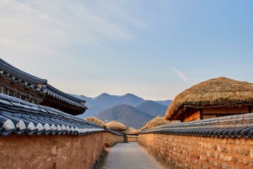 A narrow pathway lined with traditional Korean houses featuring tiled roofs and straw-covered tops, set against a backdrop of hazy mountains and a serene sky. The warm, earthy tones of the walls and roofs contrast with the cool hues of the distant mountains.