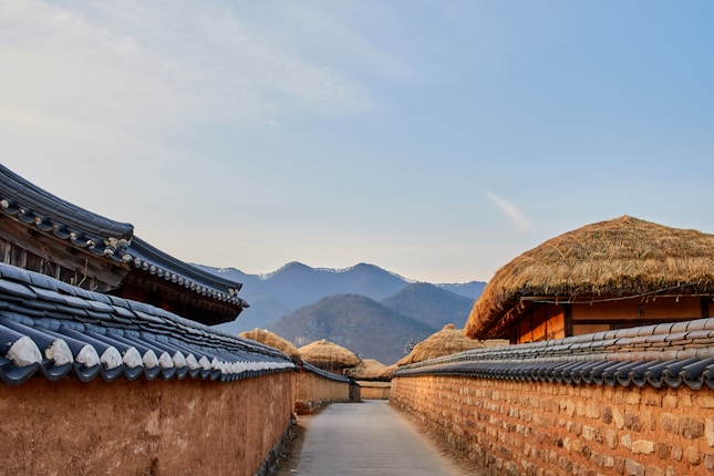 A narrow pathway lined with traditional Korean houses featuring tiled roofs and straw-covered tops, set against a backdrop of hazy mountains and a serene sky. The warm, earthy tones of the walls and roofs contrast with the cool hues of the distant mountains.