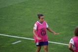A soccer player is on a field wearing a pink training vest over a white and purple uniform. The player appears to be moving with focus and concentration. The grass is green and well-maintained.