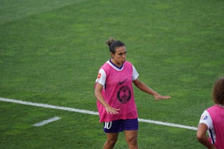 A soccer player is on a field wearing a pink training vest over a white and purple uniform. The player appears to be moving with focus and concentration. The grass is green and well-maintained.